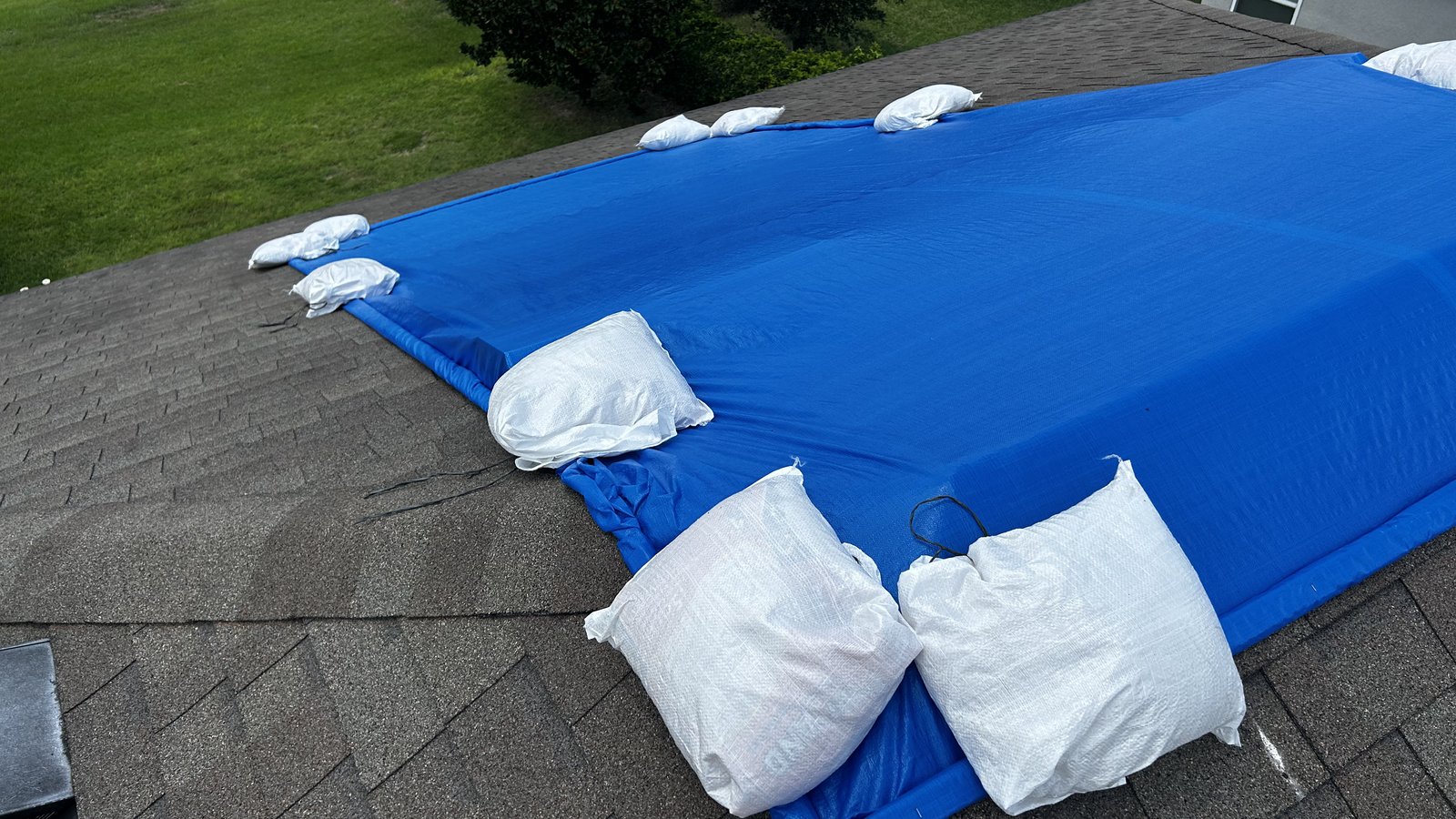 Blue emergency roof tarp held down by white sandbags along its edges on a gray shingle roof